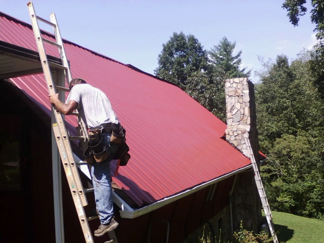 Roofer on a ladder beside a red metal roof and stone chimney — Soffit & Fascia Repair by Roofing Brothers in Marietta